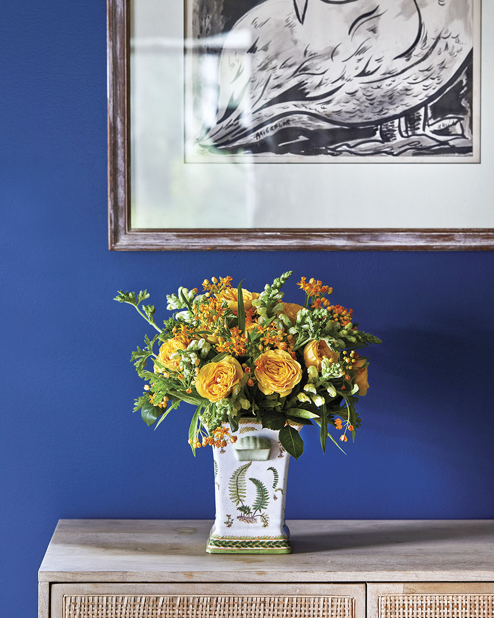 A vibrant yellow floral arrangement of dianthus, roses, snapdragons, scented geranium, and asclepia (milkweed) contrasts with the royal blue wall.