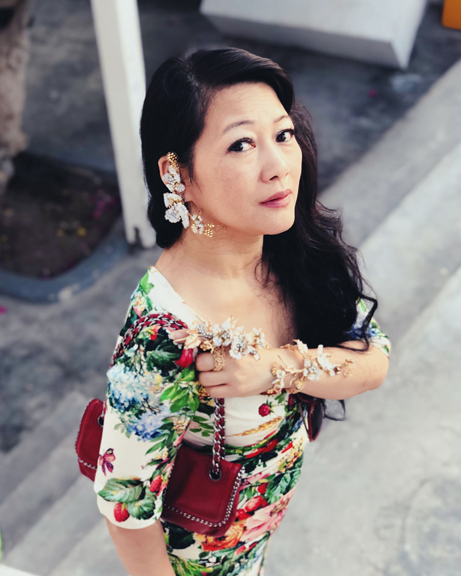 A portrait of jewelry designer Mindy Lam, a woman of asian descent with long wavy black hair, wearing a floral dress and her floral-inspired ring, bracelet cuff and earrings. She holds red clutch tucked under one arm.