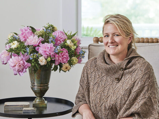 Portrait of floral designer Kirk Whitfield, wearing dark jeans and a tan cable knit shawl-style sweater, sitting next to a vase of pink peonies accented with smaller blue and white flowers