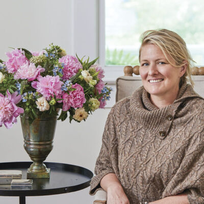 Portrait of floral designer Kirk Whitfield, wearing dark jeans and a tan cable knit shawl-style sweater, sitting next to a vase of pink peonies accented with smaller blue and white flowers