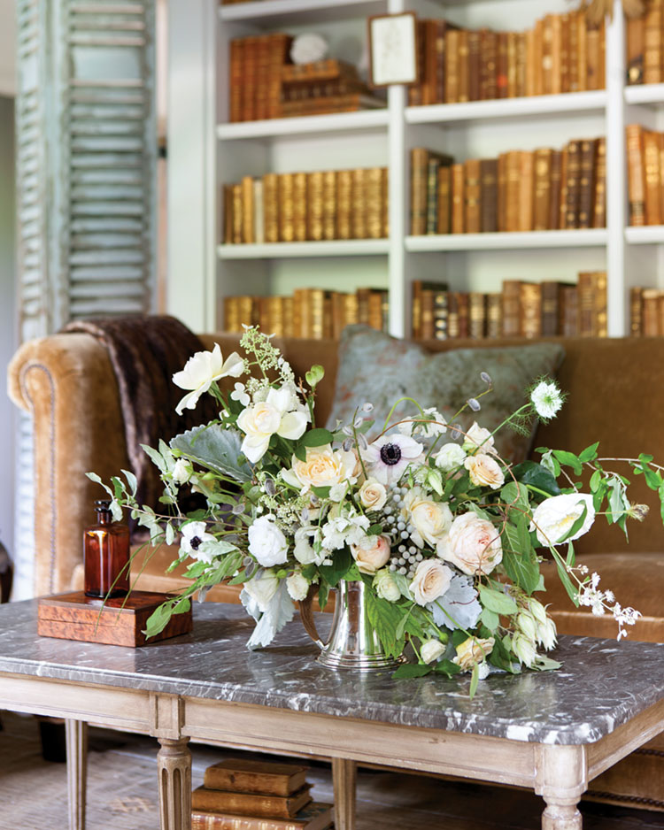 A white and green floral design by Amy Osaba sits a coffee table. A bronze-colored velvet sofa and floor-to-ceiling bookshelves are in the background.