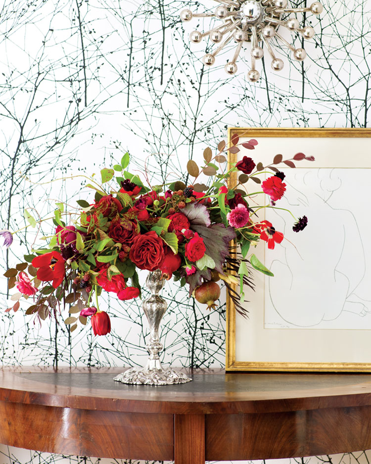 Red floral design by Amy Osaba in a tall pedestal silver container stands on demi-lune side table. The natural wood tones of the table contrast against the stark, modern wallpaper featuring motifs of bare winter trees in black on a stark white background.