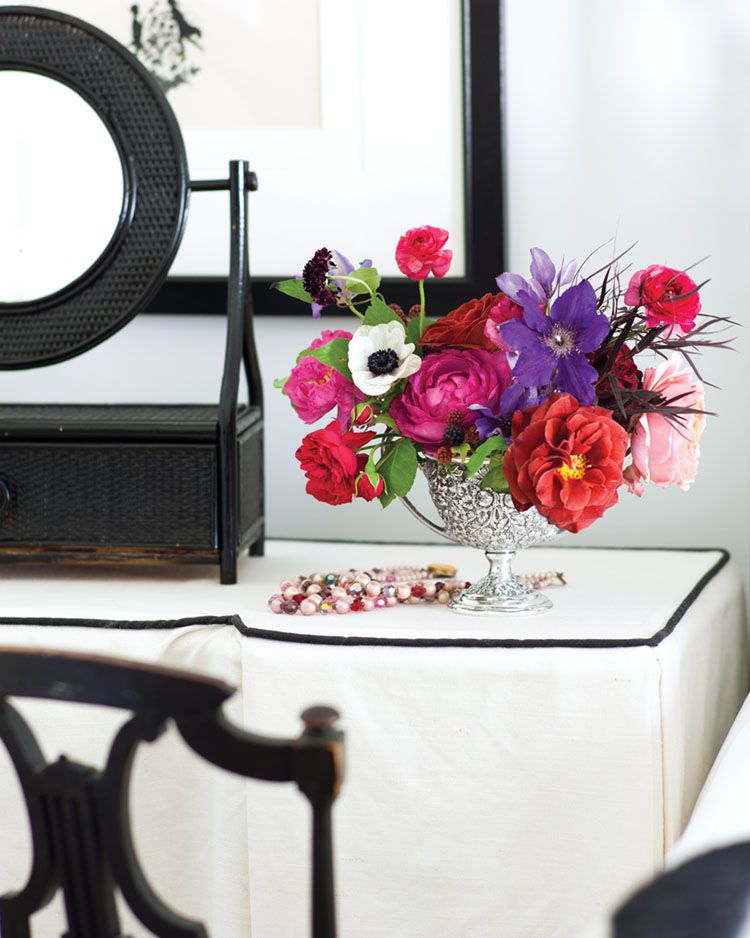 Floral design by Amy Osaba featuring bright pink, purple and red blooms in an antique silver vessel. The vase stands beside a pile of colorful jewelry on a white-skirted vanity. White walls contrast against ebony-stained chair and mirror.