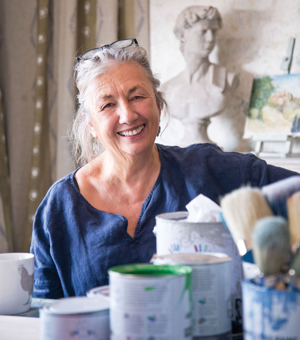 portrait of Annie Sloan with cans of her chalk paint. A bust and a painting on an easel are visible in the background. Among the colors is a tin of Amsterdam Green, a vibrant Kelly Green color