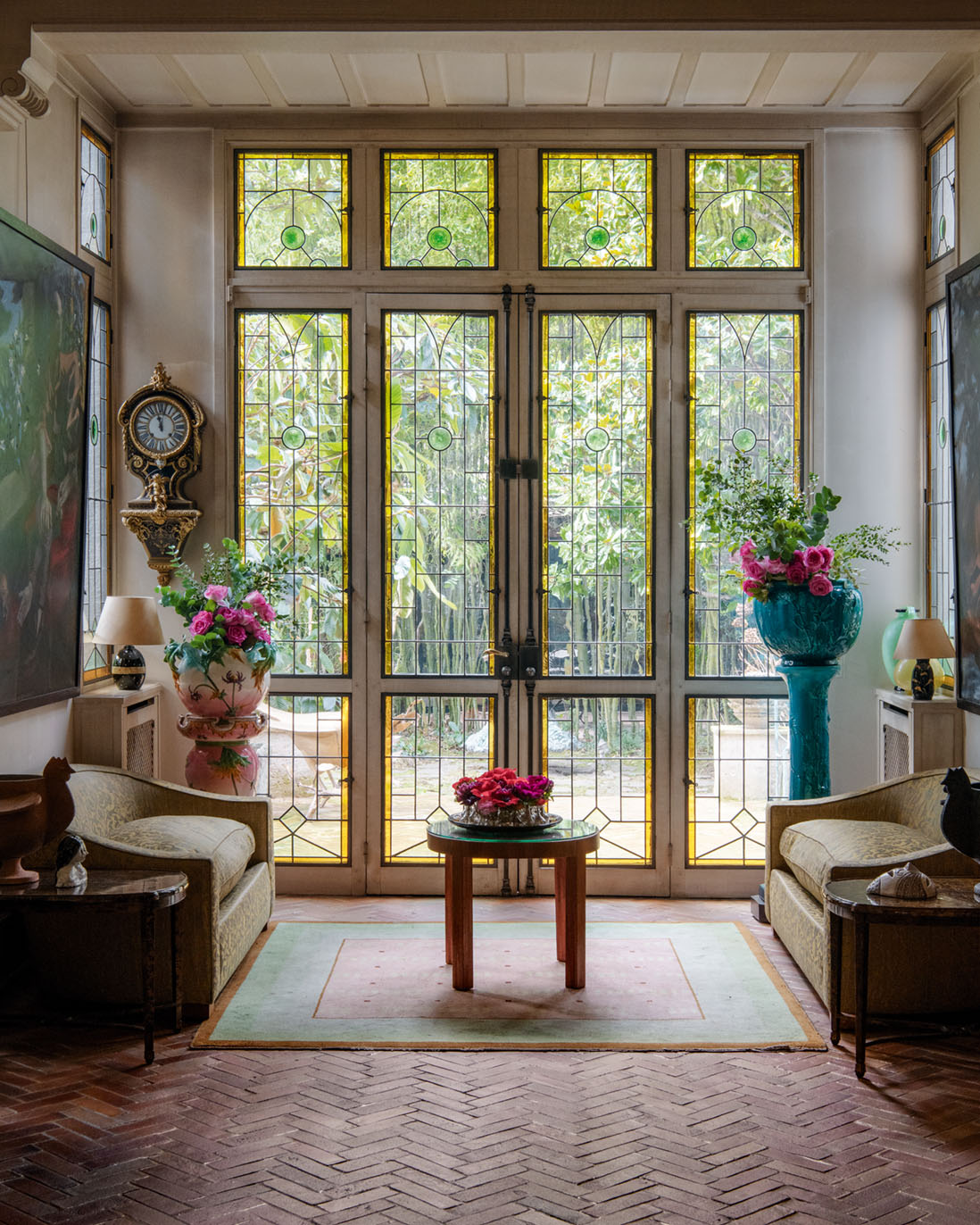 A scene from the Parisian home of Terry De Gunzburg featuring a floor to ceiling leaded glass window, a brick floor laid in a herring bone pattern, two cozy love seats facing each other, and bright pink flower arrangements placed on the coffee table, and on pedestals flanking the window.