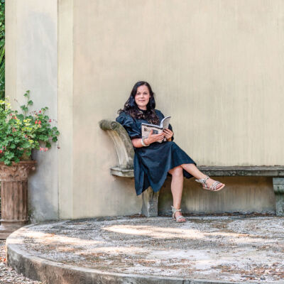 Betty Soldi, author of Inkspired (translated into eight languages), sits on a sandstone bench leaning against a wall decorated with classical flourishes.
