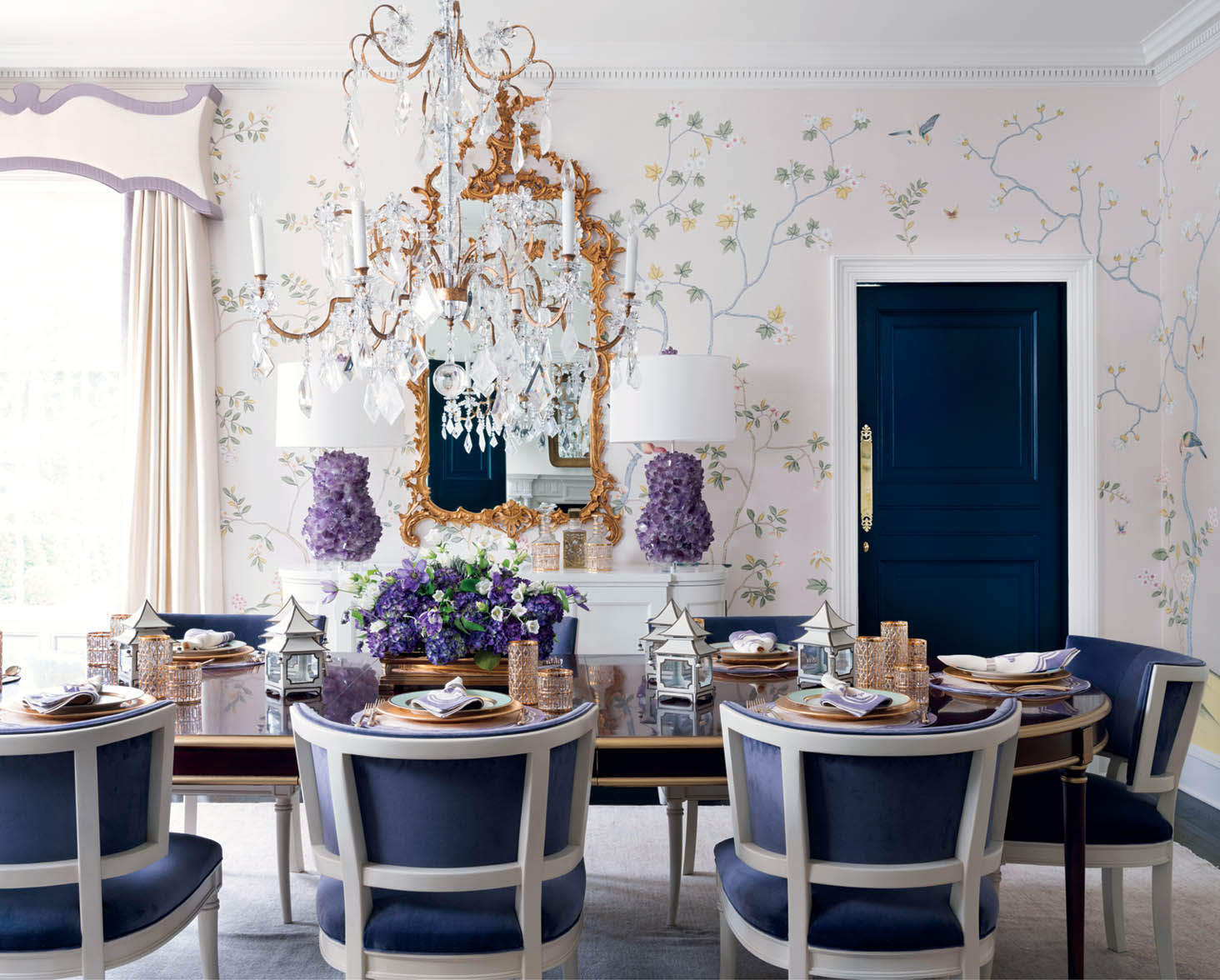 A dining room by interior designer Melanie Turner, featured in her book, Inviting Interiors. The room features a light and airy floral wallpaper on a white background, dental molding, a grand crystal chandelier, a gilt mirror flanked by two lamps with amethyst bases. She mixes wood tones, with a dark wood dining table with gild edges pared with white-wood framed dining chairs with navy upholstery. The door is painted navy.