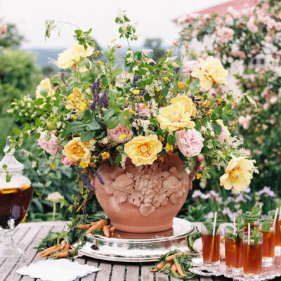 A large yellow, pink, and green floral arrangement at the center of a table filled with glasses of sweet tea, table set in the rose garden at Moss Mountain Farm, created by The Velvet Boxwood for duck, duck, goose-themed children's social