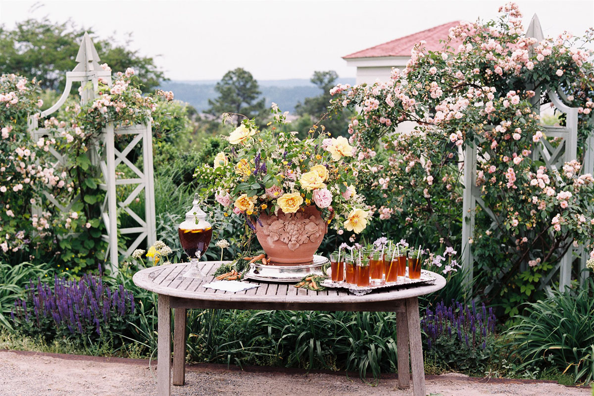A large spring floral arrangement on a drinks table in the garden. Rose-covered tuteurs bloom in the background