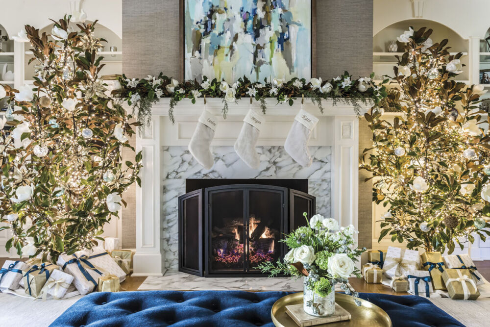 A pair of magnolia Christmas trees flank a mantel decorated with a garland and white stockings for the holidays in a Raleigh, NC, home decorated by Vicky Serany