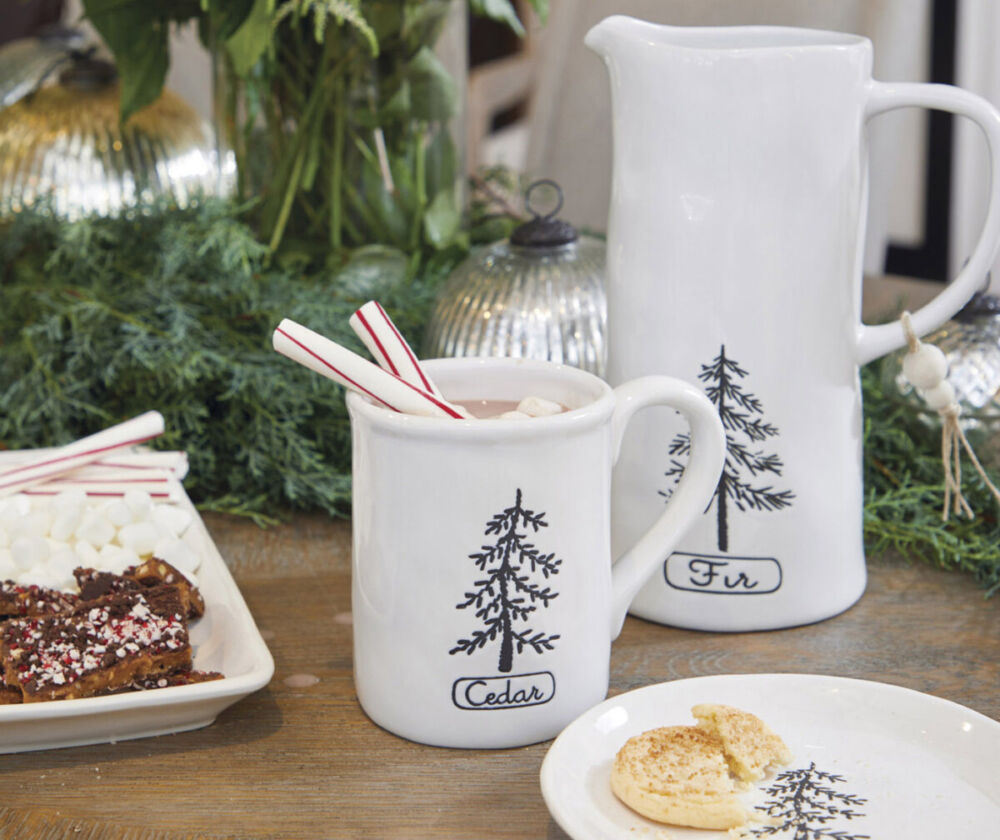 A pattern bearing an illustration of a simple, singular evergreen tree decorates white ceramic pitchers, mug, plate and tray from Summer Classics home stores. The pitcher and mug are filled with hot chocolate, and the tray and play hold cookies, soft peppermint sticks, and marshmallows on a table set for a holiday hot cocoa party.