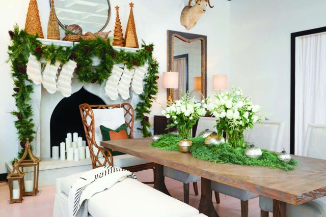 Photo of a dining room furnished with long wooden dining table, a pair of long wood benches on either side, and rattan-backed chairs at either end. A wreath, a long green garland, and small Christmas tree-shaped decorations decorate a white plaster mantel with an arabesque opening for the fire. The table is decorated with garlands, mercury glass votives, and vases of white flowers. The furniture and holiday decor is sold at Summer Classics Home stores.