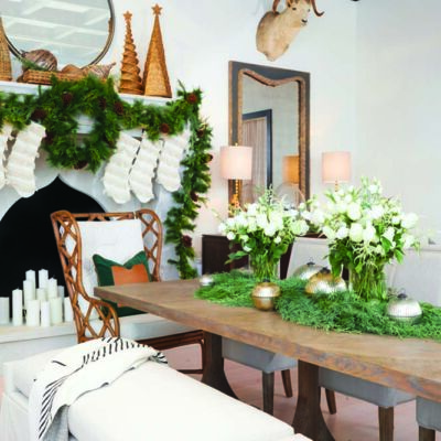 Photo of a dining room furnished with long wooden dining table, a pair of long wood benches on either side, and rattan-backed chairs at either end. A wreath, a long green garland, and small Christmas tree-shaped decorations decorate a white plaster mantel with an arabesque opening for the fire. The table is decorated with garlands, mercury glass votives, and vases of white flowers. The furniture and holiday decor is sold at Summer Classics Home stores.