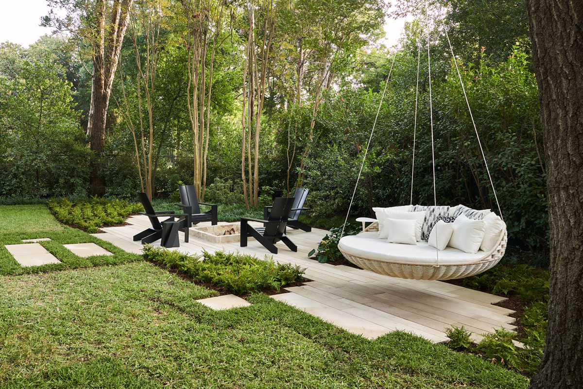 4 adirondack surround a firepit on a backyard patio. A daybed swing hangs from a tree