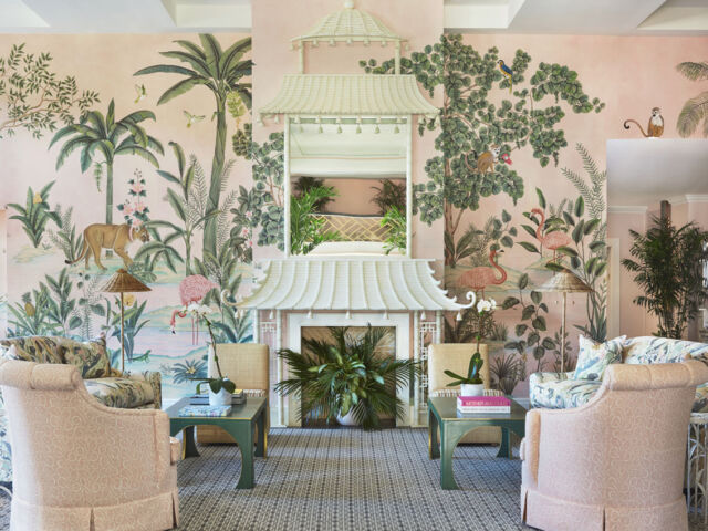 Central seating area in the lobby of the Colony Hotel in Palm Beach, featuring custom scenic wallpaper de Gournay Wallpaper from the lobby’s 2021 renovation by Kemble Interiors. The wallpaper depicts flora and fauna native to the area. A symmetrical seating arrangement flanks a white fireplace mantel made to look like a pagoda. The room’s color palette includes soft pinks and greens.