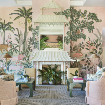 Central seating area in the lobby of the Colony Hotel in Palm Beach, featuring custom scenic wallpaper de Gournay Wallpaper from the lobby’s 2021 renovation by Kemble Interiors. The wallpaper depicts flora and fauna native to the area. A symmetrical seating arrangement flanks a white fireplace mantel made to look like a pagoda. The room’s color palette includes soft pinks and greens.