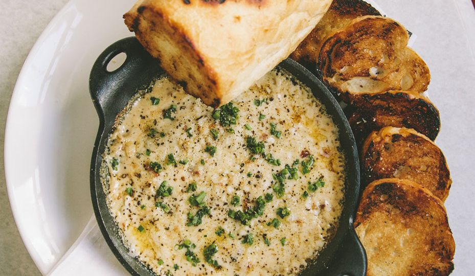 Artichoke Dip in a cast-iron pan, on a platter surrounded by grilled baguette slices
