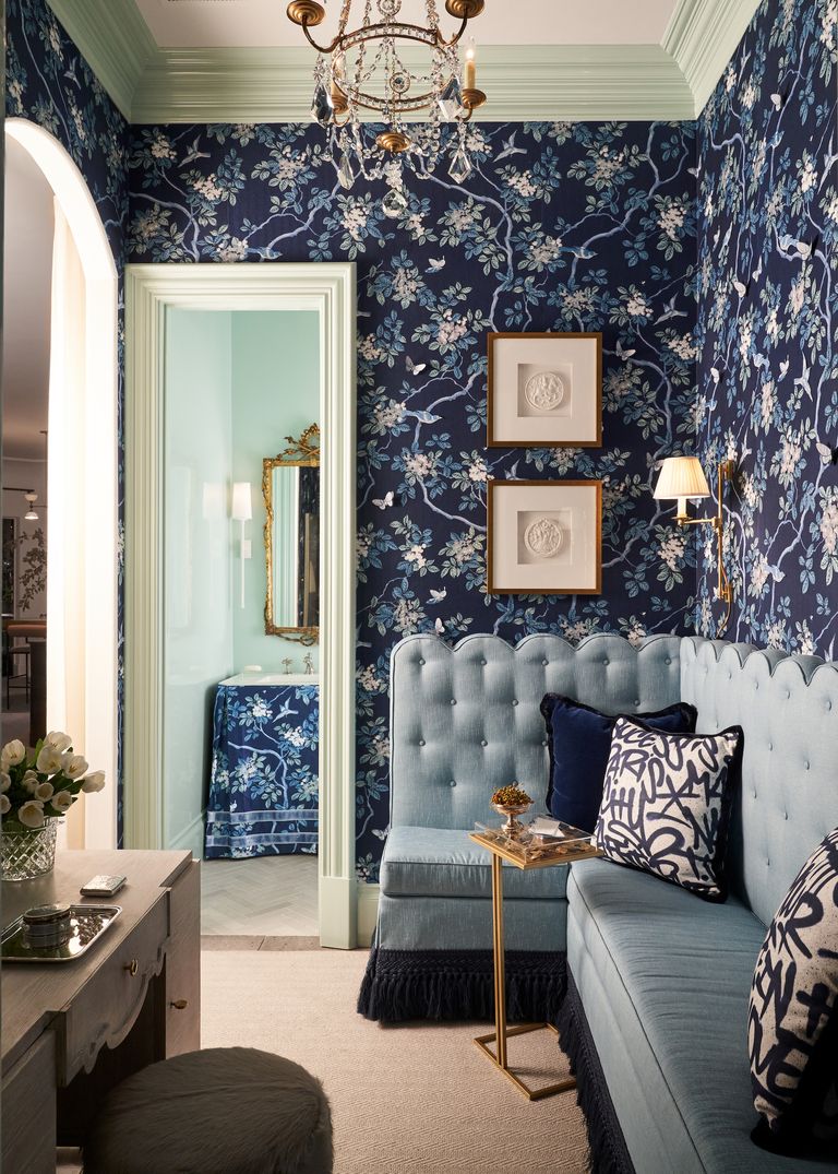 In a space designed by Traci Zeller for the Kips Bay Dallas Showhouse 2020, a dark blue floral print covers the walls of a sitting room and skirts the sink in the powder room beyond, where the walls are a pale blue. The sitting room also features a large, pale blue tufted banquette.
