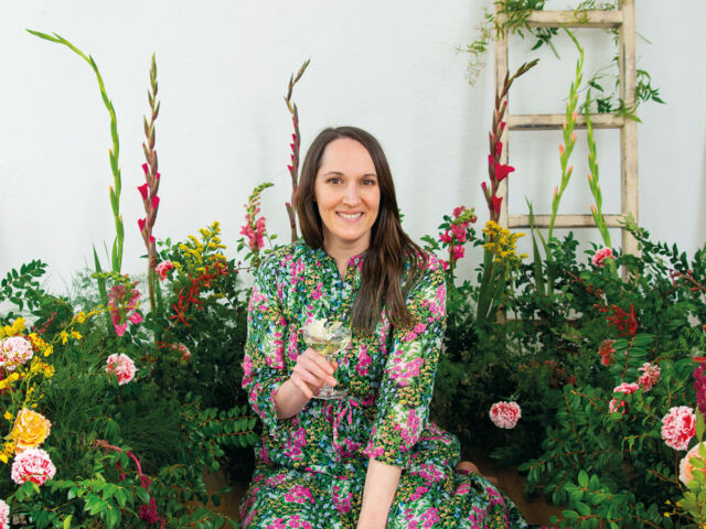 portrait of Alyson Brown, author of The Floral-Infused Cocktail, wearing a floral dress, surrounded by a lush floral installation, holding a cocktail in her hand