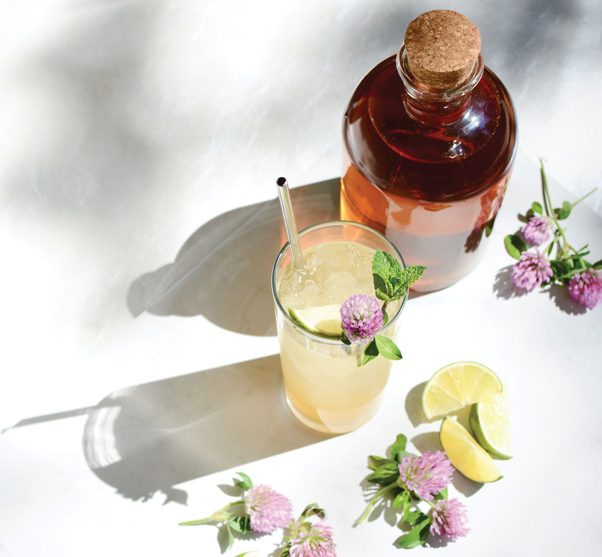 photo of a bottle of Clover Sun Tea, and a glass of the Cloverjito cocktail, garnished with lime wedges and purple clover flowers