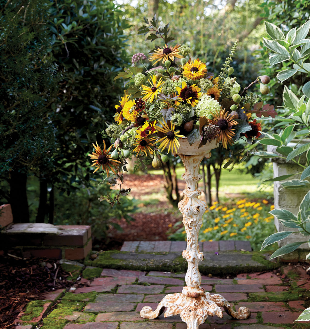 summer flower arrangement with yellow, rust, and green towns, arranged on an antique wrought iron pedestal piece painted white with rust showing. The pedestal stands on a mossy brick patio, with a lush summer garden in the background.