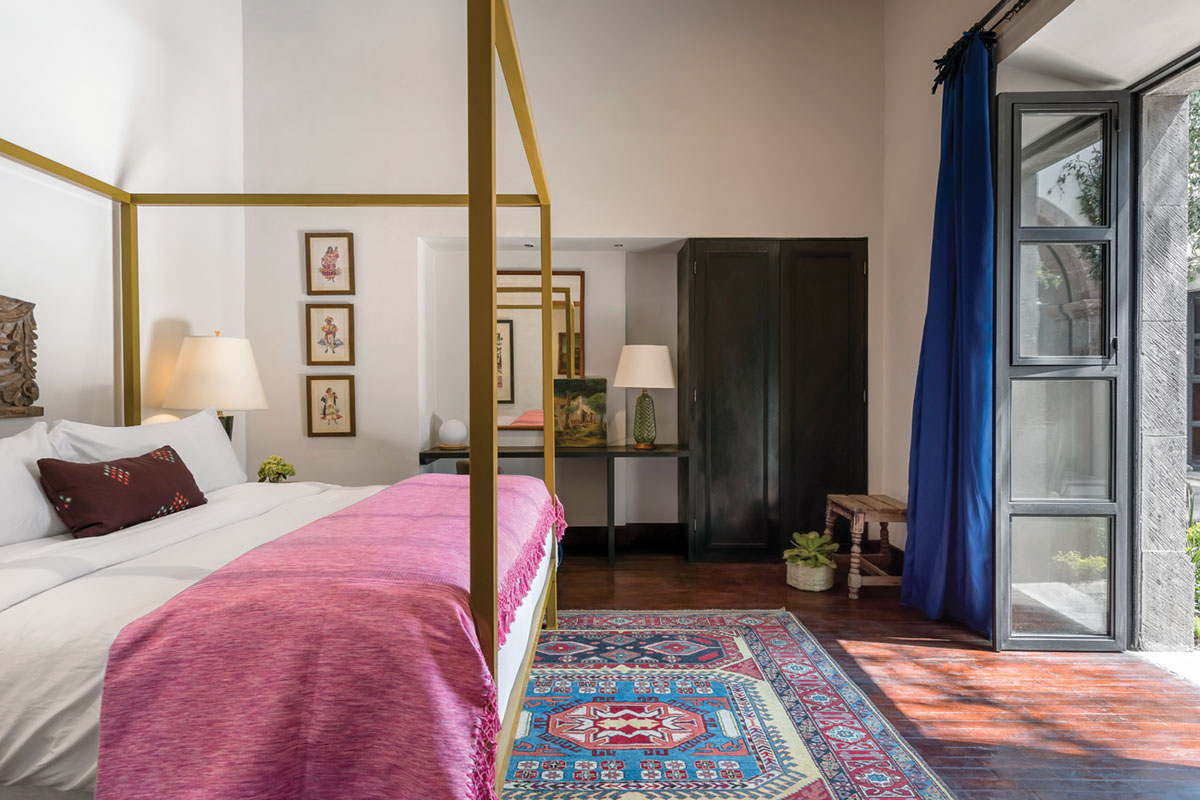 guest room with a white stucco walls, high ceilings, tapestry rug on wood floors, cheerful dark pink bedspread on an uncovered canopy-frame bed, and large french doors framed by royal blue drapery panels leading outside. The doors are open to let in the pleasant climate of San Miguel de Allende, Mexico