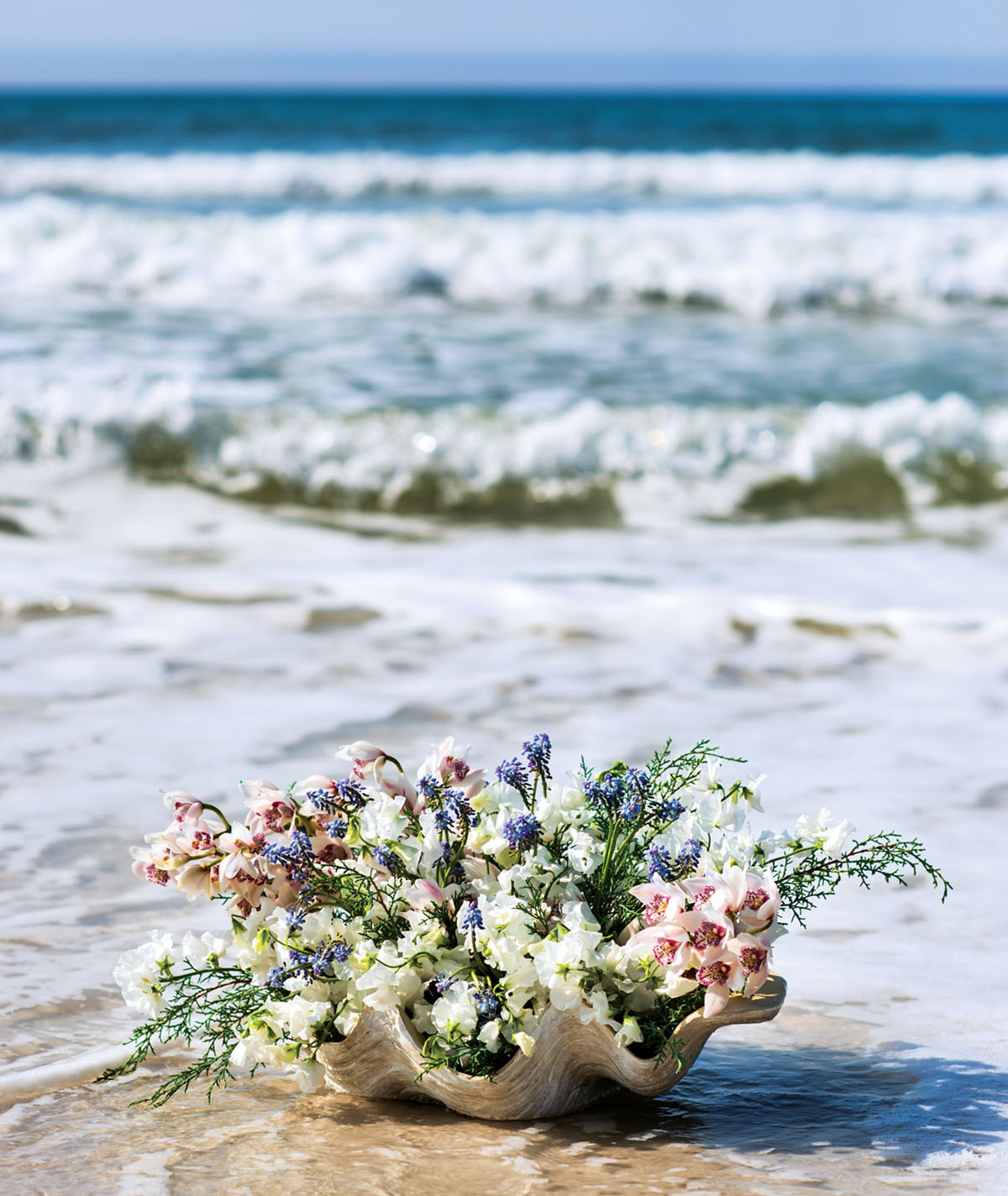 large half shell filled with flowers set on the shore with waves and blue sky in the background