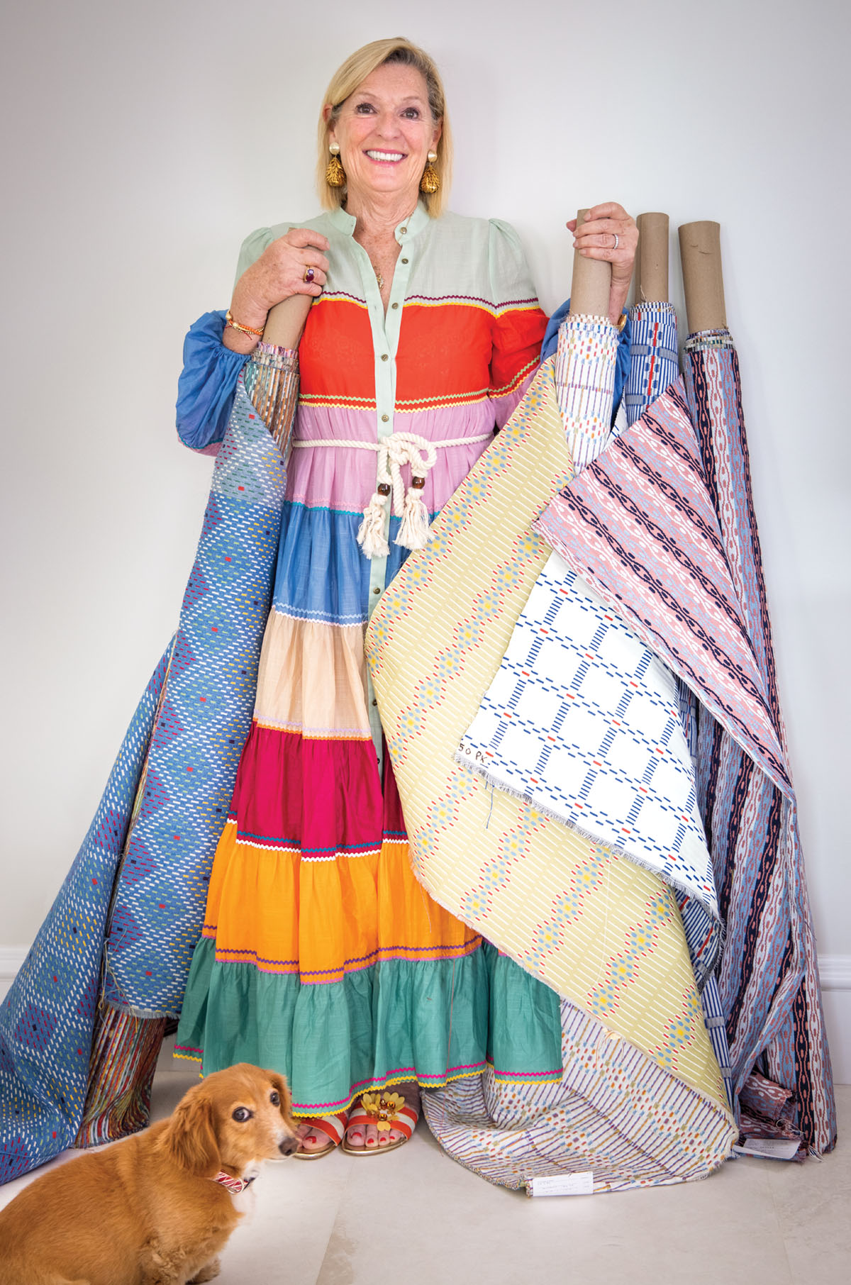 Portrait of interior designer Mally Skok wearing a long flowy dress with wide multi-color stripes, holding bolts of fabric from her collection. A small dog stands at her feet
