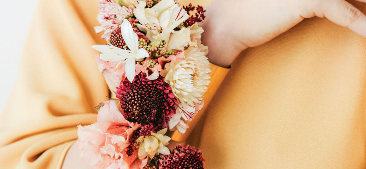 A model wears a floral cuff on her wrist featuring burgundy, cream, and peach-colored fresh flowers