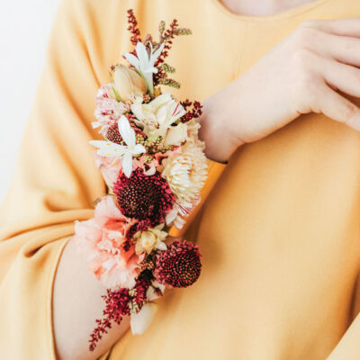 A model wears a floral cuff on her wrist featuring burgundy, cream, and peach-colored fresh flowers