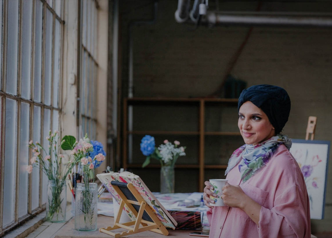 British artist Safiyyah Choycha standing by a window in her studio, wearing a turbin, floral neck scarf, and pink artist smock