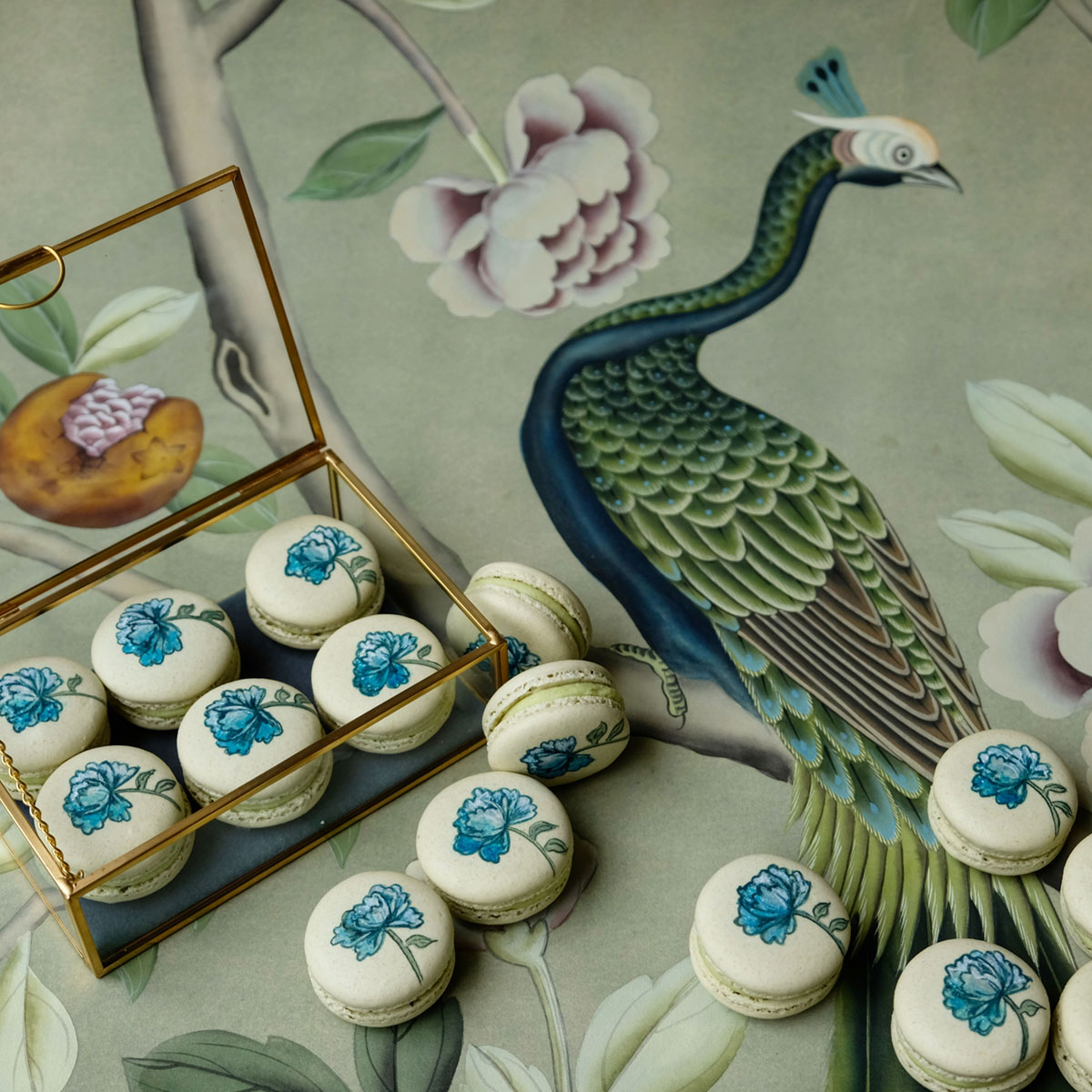 Macarons featuring a blue-and-white floral design are displayed in a glass box on a fabric pattern featuring birds and botanical elements