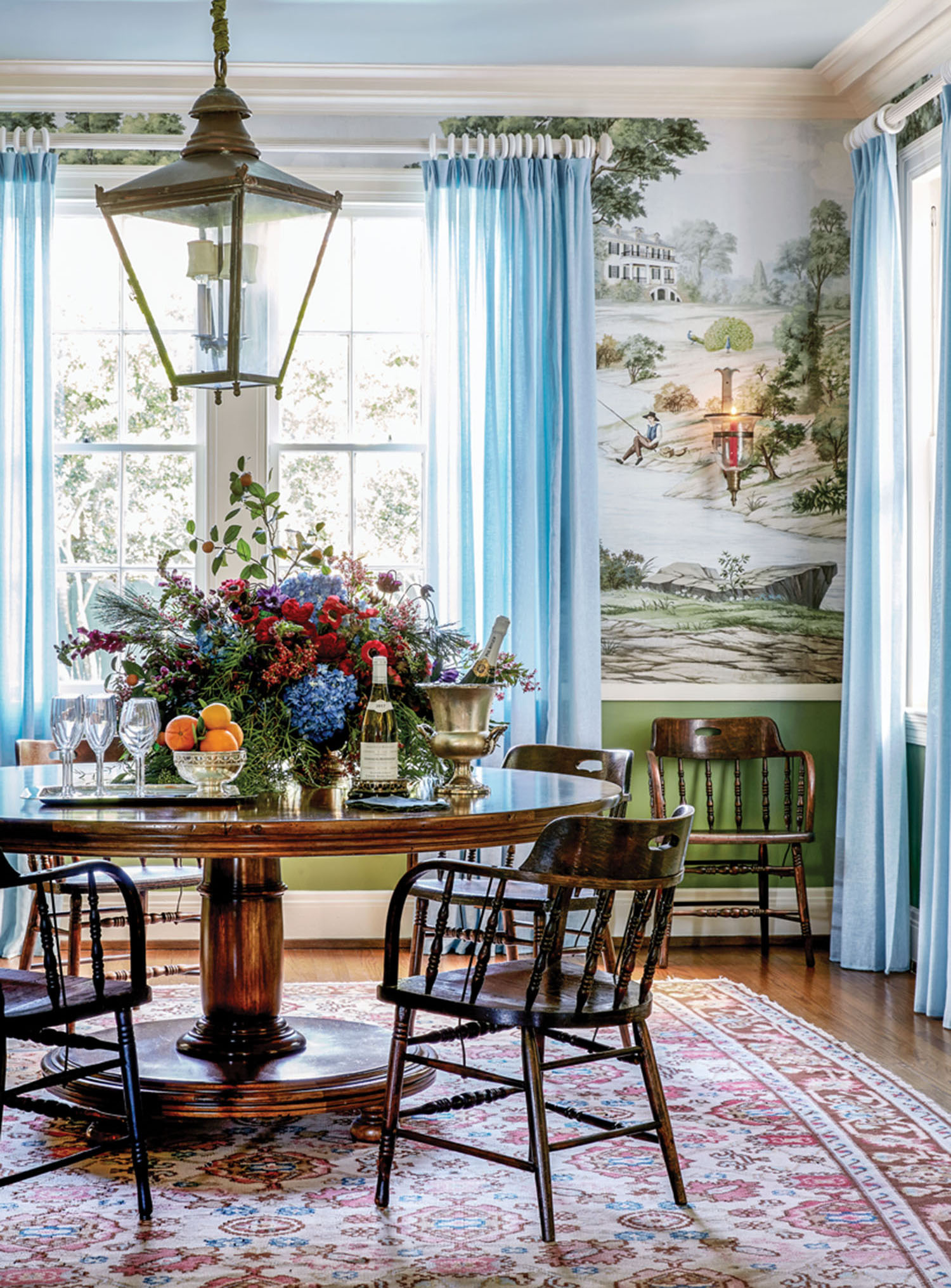 Dining room in the New Orleans home of Ware Porter. Features: Scenic wallpaper in a green colorway, pale blue sheer drapery panels, antique circular wood dining table and chairs, oriental rug, pale blue ceiling