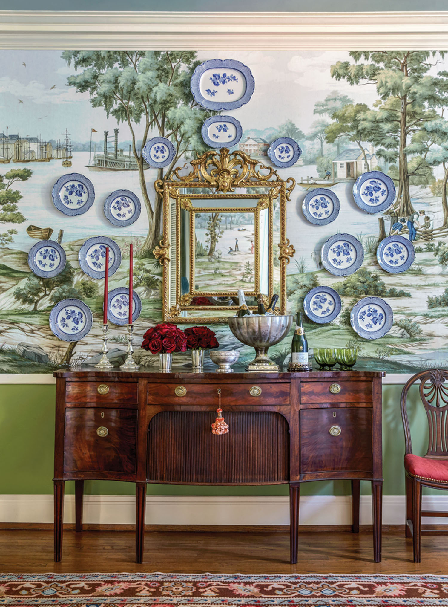 A collection of blue-and-white plates is displayed on a wall covered in scenic wallpaper in a green colorway, above an antique wooden sideboard. Laid out on the sideboard are a pair of tall thin burgundy tapers in silver candleholders, lush burgundy flowers in a pair of silver tumblers, champagne bottles chilling in a silver pedestaled champagne bucket, and green drinking glasses