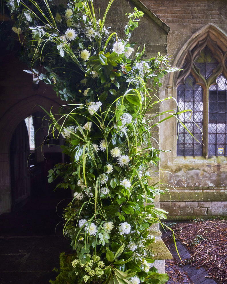 naturalistic floral arch with lush greenery and white flowers. In the background: the stone facade and leaded window of an old English church