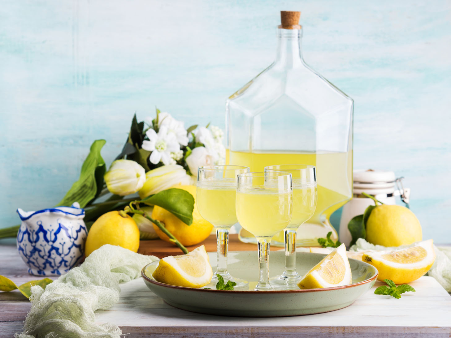 vignette with glass jug of homemade limoncello, stemmed glasses of limoncello on a tray, sliced lemons, white flowers, a canister of sugar, and a small blue-and-white porcelain vessel