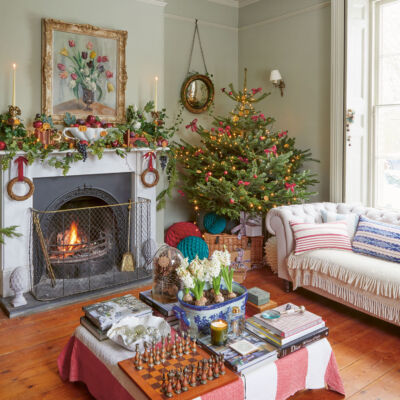 Paula Sutton’s light-filled, cozy living room at Hill House features pale green walls, a gilt-framed painting of tulips above a roaring fireplace, and a rolled-arm, tufted white sofa. On the large square coffee table, a blue-and-white dish holds blooming hyacinth bulbs. Covered with a faded vintage quilt with wide red and white stripes, the coffee table also holds books, a chess board, and a glass dome filled with pinecones. The mantel is decorated for the holiday season with greenery, grapes, apples, and dried orange slices. In the corner is a petite but full Christmas tree decorated with red bows and dried orange slices.
