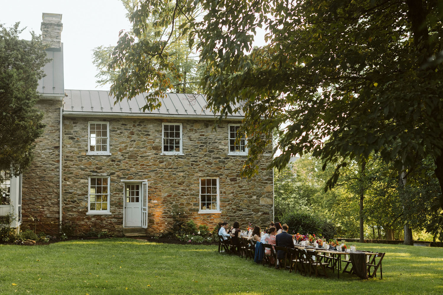A two-story stone farmhouse with a chimney. A long table is set on the lawn near the shade of a tree
