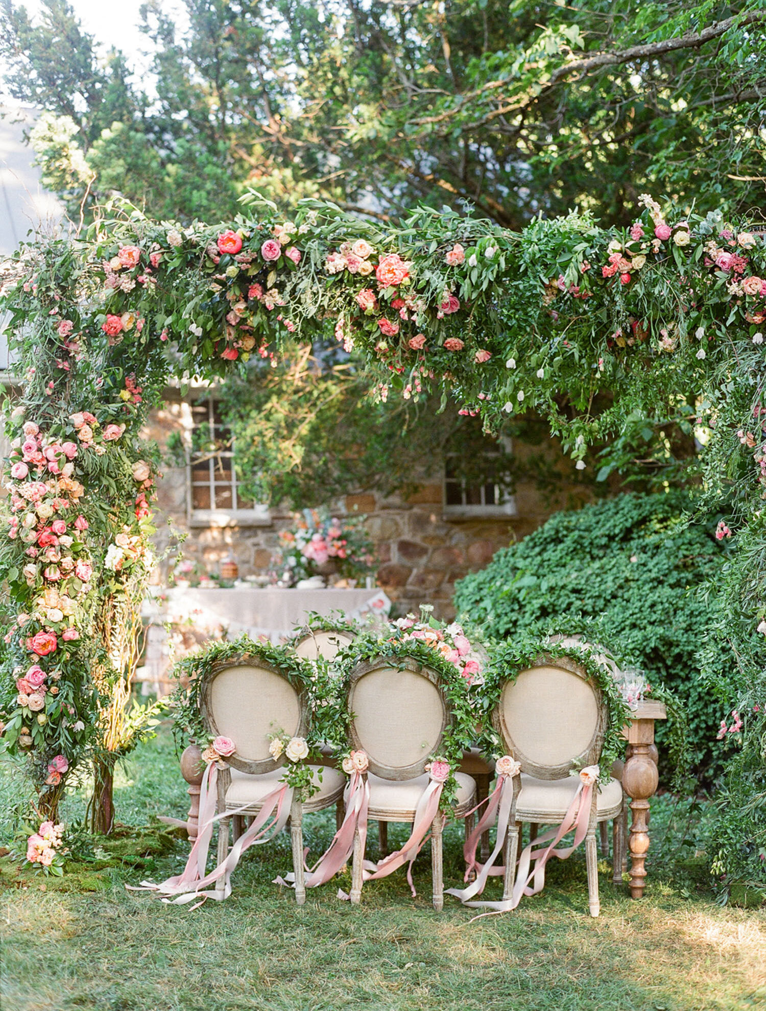 An outdoor table for six is set under a lush floral arch by Holly Heider Chapple Flowers. The rounded chair backs are trimmed in greenery with long flowing pink ribbons on each end