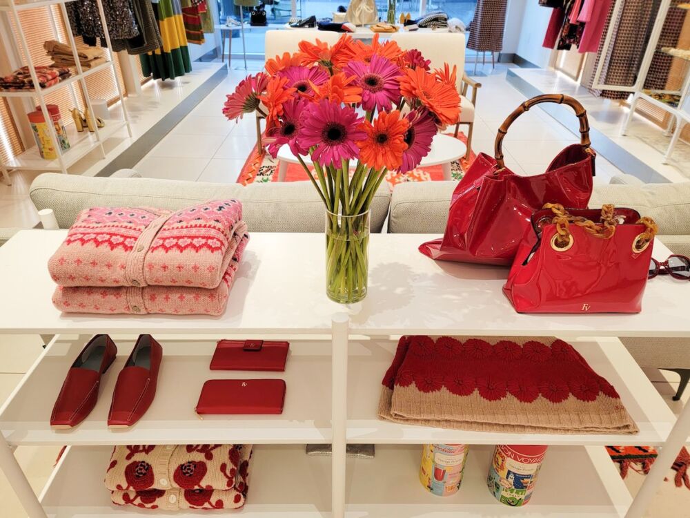 Vase of pink and orange gerberas on shelf displaying sweaters, bags, shoes in shades of red.