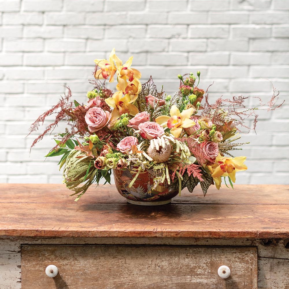 Textural arrangement of roses, cymbidium orchids and other flowers in pinks and yellows.