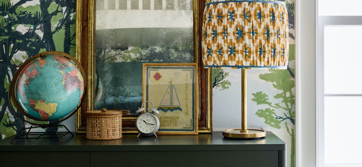 Chest topped with globe, mirror, and lamp with drum shade in boys' bedroom. Woodland wallpaper in blues and greens on wall.