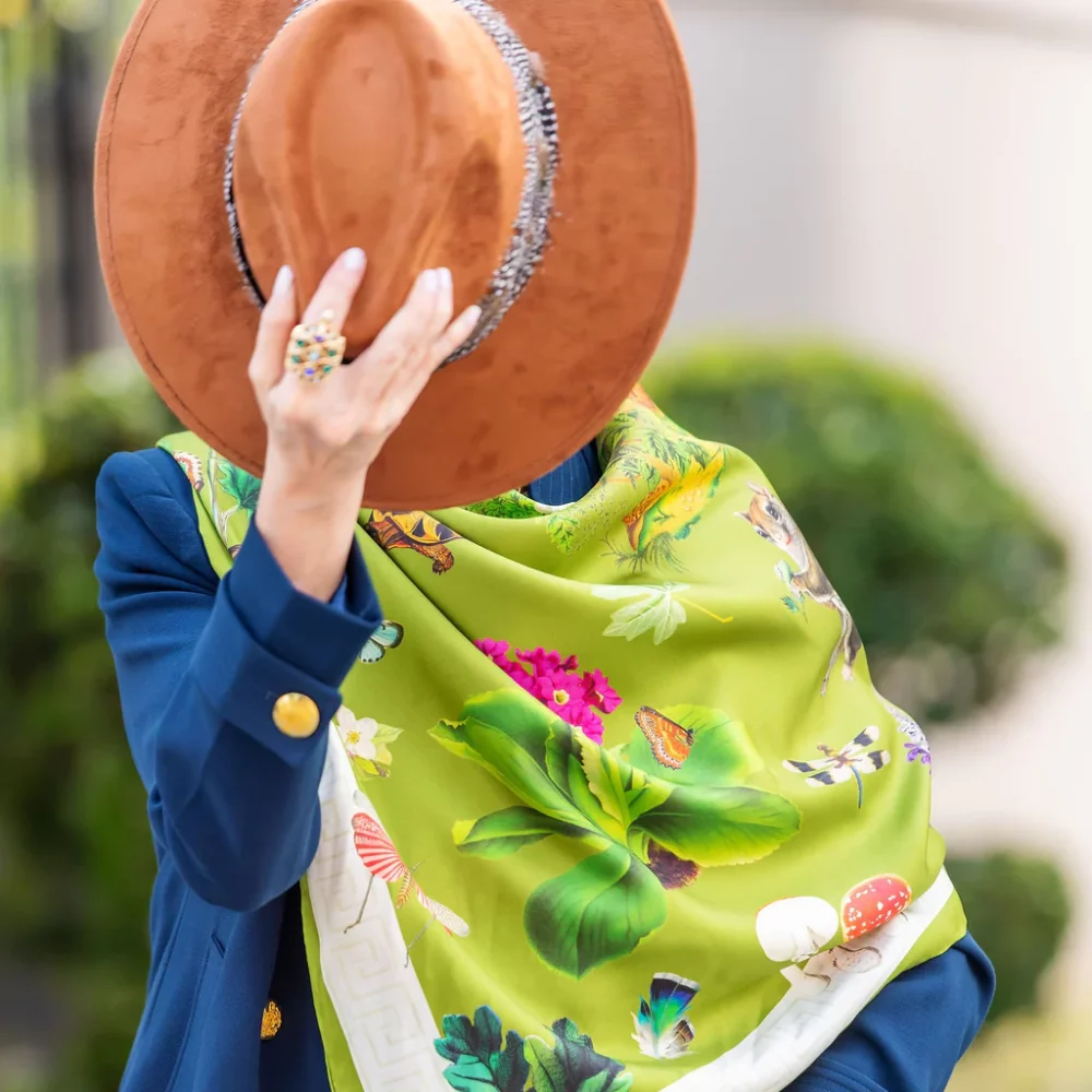 A woman in a cobalt jacket dons a bright lime green scarf with colorful bugs and plants on it.
