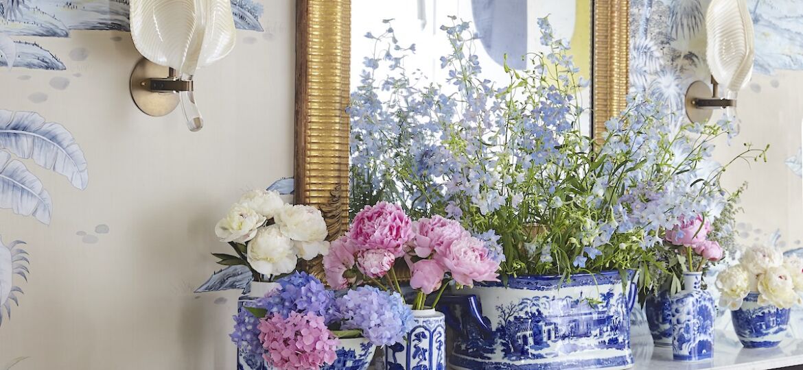 Chest with blue and white China vessels filled with arrangements of peonies, larkspur, and hydrangeas. A pair of leaf-shaped wall sconces flank a gold-framed mirror over the chest.