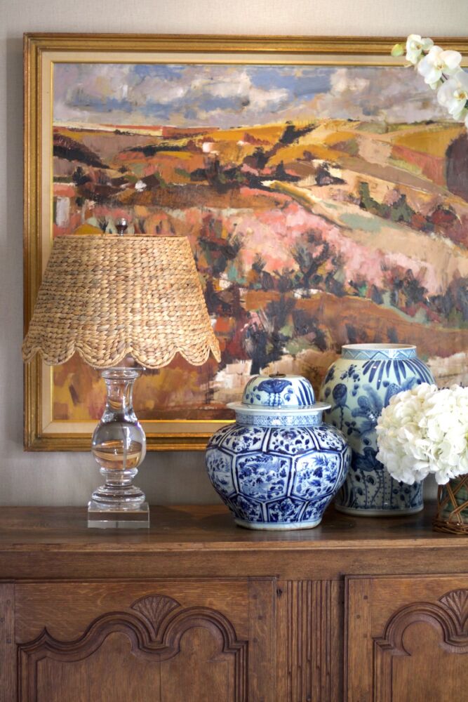 Empire, woven water hyacinth lampshade on a clear glass lamp atop wooden chest with blue and white jar and vase.