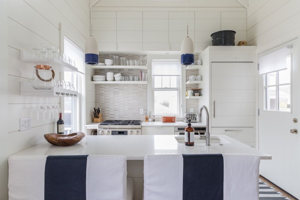 Kitchen with white shiplap walls and high ceiling, ceramic pendants over peninsula, recessed light over sink.