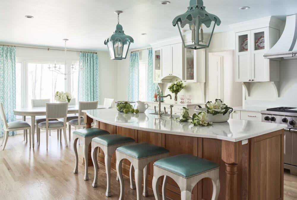 Verdigris lanterns hanging over kitchen island with blue-green upholstered barstools. White chandelier over breakfast table and aqua curtains on windows.