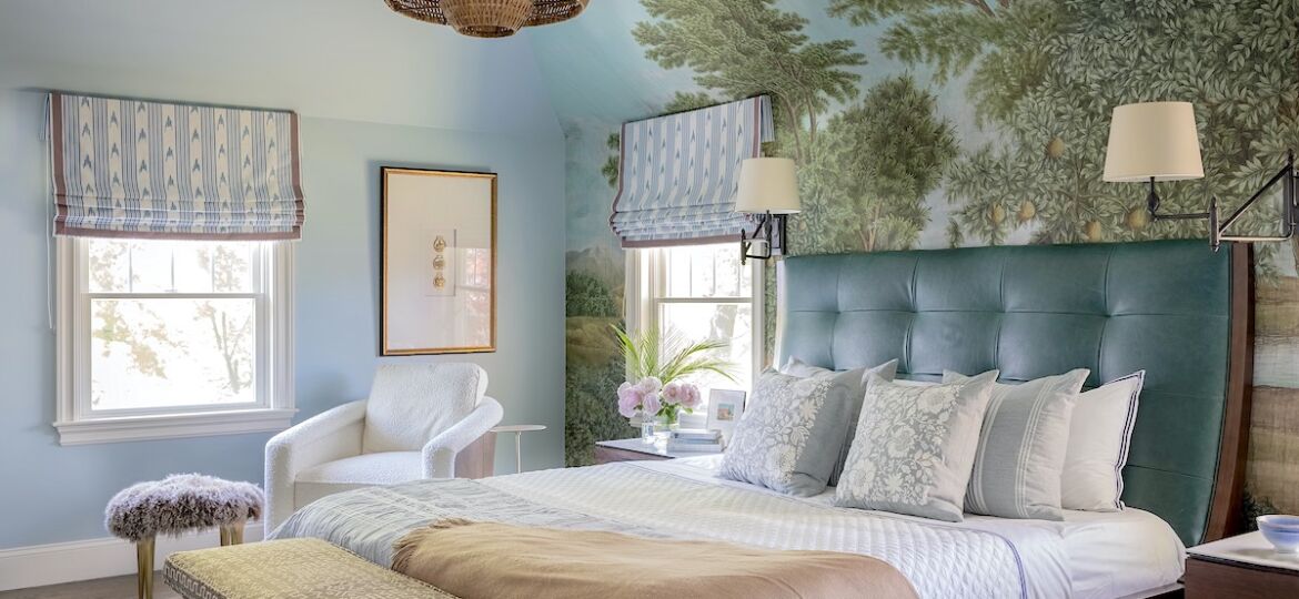 A leather headboard sits over scenic wallpaper along with a bold tired chandelier and swing arm lamps.