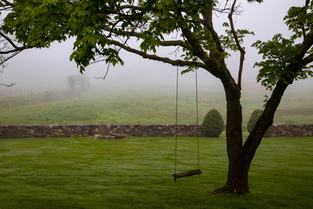 An empty wooden swing hangs from a tree in front of a stone wall and grassy meadow.