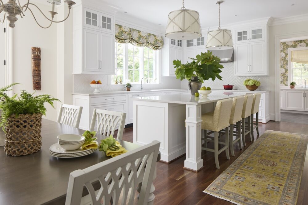 Kitchen with a fresh yellow, green, and white palette and pleated Visual Comfort pendants with brass accents. A Thibaut botanical print perks up the window over the sink. Vase of oakleaf hydrangea foliage on island and orchids with napkins on table.